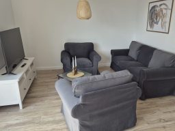 Storchennest Wohnbereich Wohnzimmer mit zwei grauen Sesseln, einer hellen Couch, TV und neutraler Dekoration.